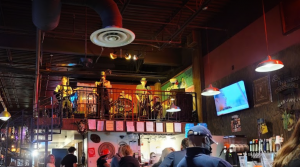 Jazz - A Louisiana Kitchen A band performs on an elevated stage in a dimly lit bar, with patrons seated below. A TV screen displays a show in the background. Kansas City, Columbia