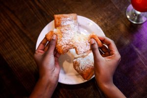 Jazz - A Louisiana Kitchen Hands pulling apart a beignet dusted with powdered sugar over a plate on a wooden table. Kansas City, Columbia