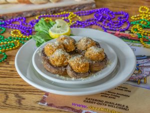 Jazz - A Louisiana Kitchen A plate of stuffed mushrooms topped with cheese. The dish is garnished with lemon and greens, surrounded by colorful beads on a wooden table. Kansas City, Columbia