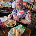 Jazz - A Louisiana Kitchen A man seated indoors, wearing a hoodie with an American flag pattern, smiles while eating a meal. A basket of bread and two small containers sits on the table in front. Kansas City, Columbia, cajun food restaurant near me