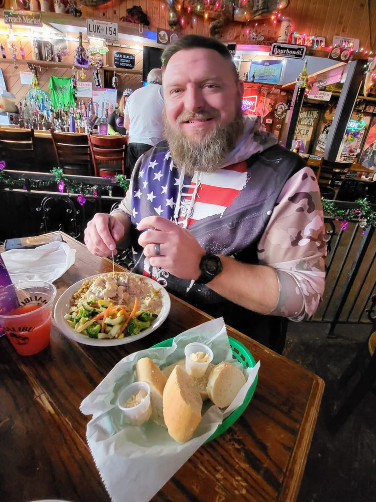 Jazz - A Louisiana Kitchen Man in a bar wearing a patriotic hoodie, smiling while eating a plate of food with vegetables and bread on the table. Kansas City, Columbia, cajun food restaurant near me