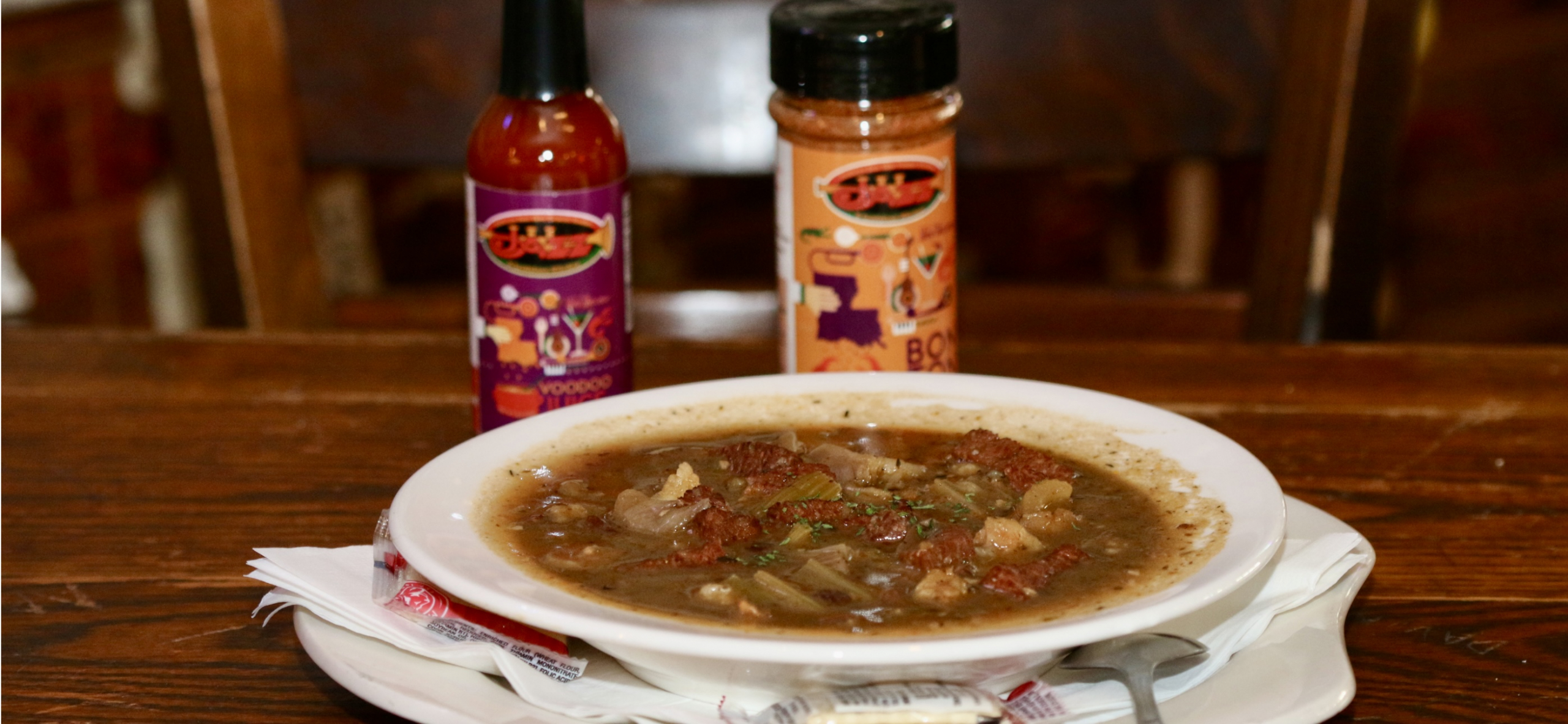 Jazz - A Louisiana Kitchen A bowl of gumbo is on a table, accompanied by a bottle of hot sauce and a seasoning shaker in the background. Napkins are placed beneath the bowl. Kansas City, Columbia, cajun food restaurant near me