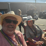 Jazz - A Louisiana Kitchen Two people wearing hats and colorful bead necklaces are sitting outdoors, smiling at the camera. Sunlight is visible in the background, with other people and buildings in the vicinity. Kansas City, Columbia, cajun food restaurant near me