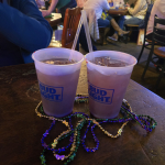 Jazz - A Louisiana Kitchen Two plastic cups with drinks and straws sit on a table, adorned with colorful Mardi Gras beads. People are conversing in the background. Kansas City, Columbia, cajun food restaurant near me