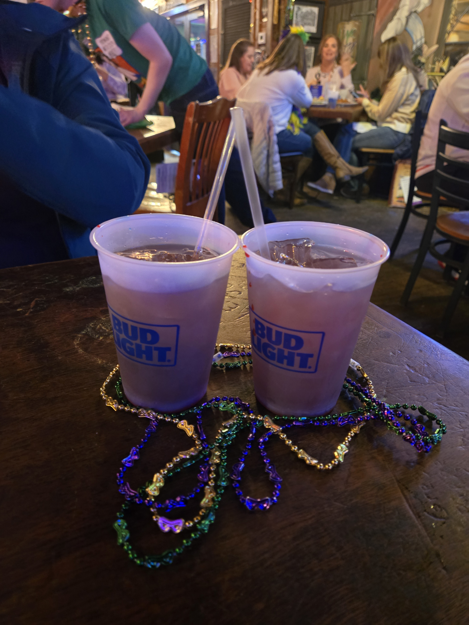 Jazz - A Louisiana Kitchen Two plastic cups with drinks and straws sit on a table, adorned with colorful Mardi Gras beads. People are conversing in the background. Kansas City, Columbia, cajun food restaurant near me