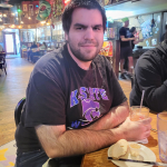 Jazz - A Louisiana Kitchen Man sitting at a wooden table in a restaurant, wearing a black K-State shirt. He is holding a drink and looking at the camera. Plates and drinks are on the table. Kansas City, Columbia, cajun food restaurant near me