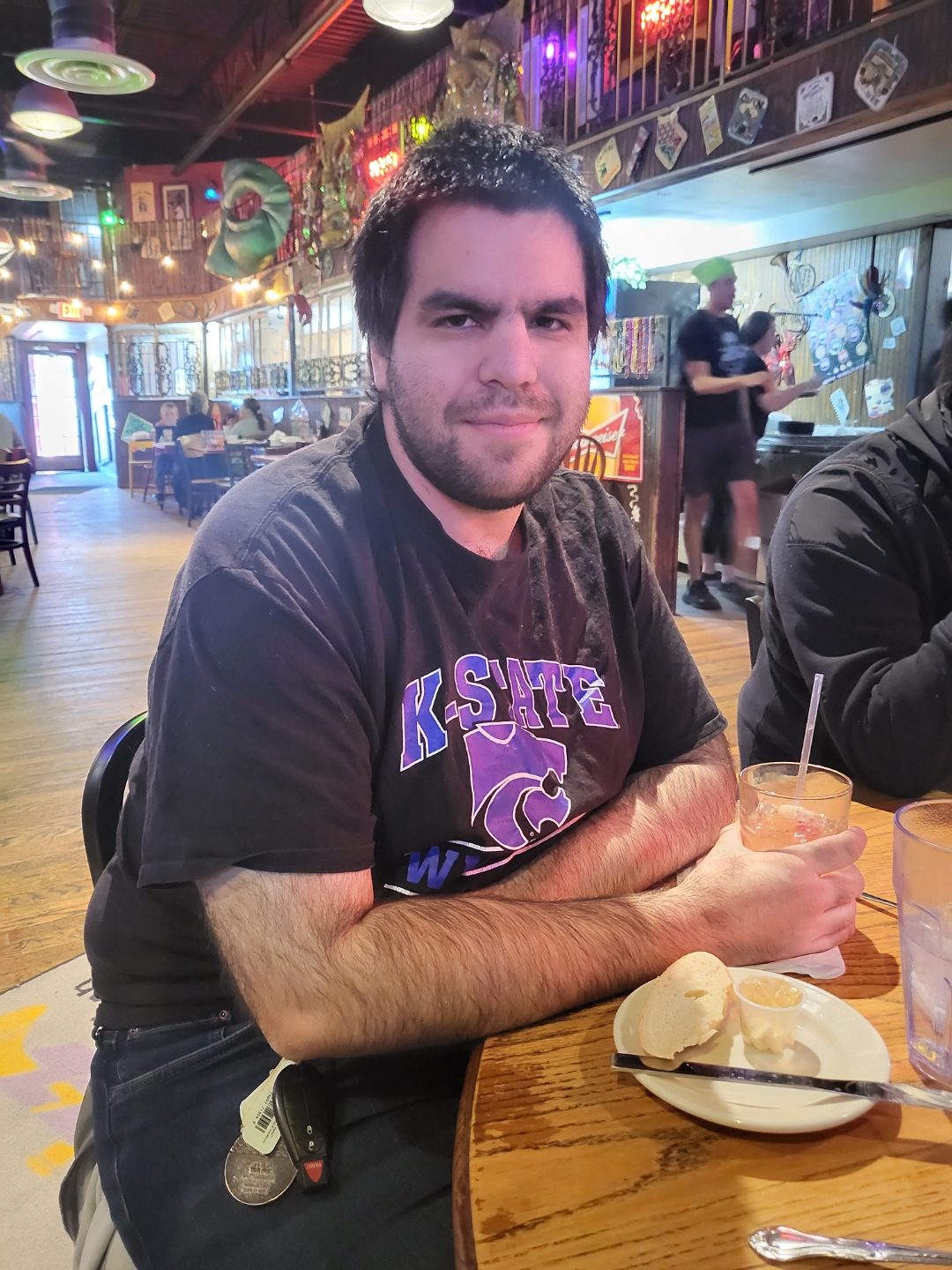 Jazz - A Louisiana Kitchen Man sitting at a wooden table in a restaurant, wearing a black K-State shirt. He is holding a drink and looking at the camera. Plates and drinks are on the table. Kansas City, Columbia, cajun food restaurant near me